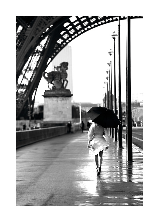  – Fotografia in bianco e nero di una donna che cammina riparandosi con un ombrello sotto la Torre Eiffel