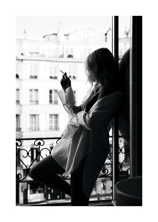  – Fotografia in bianco e nero di una donna che indossa una camicia oversize e fuma una sigaretta in piedi a una finestra