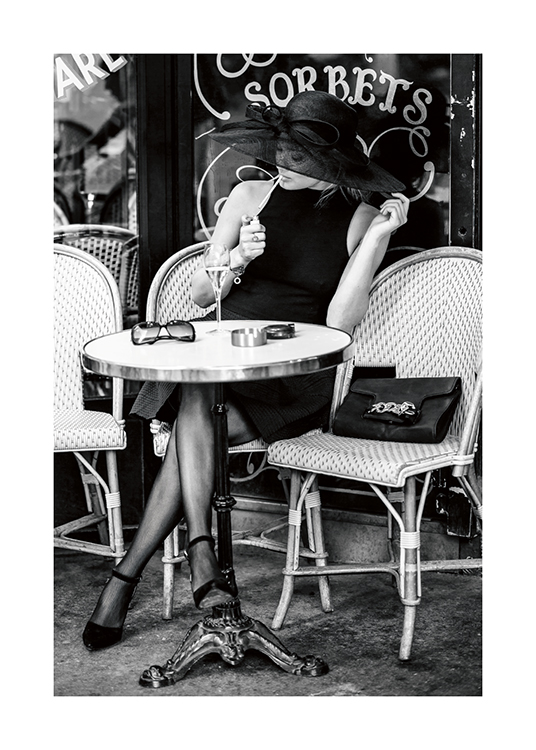  – Fotografia in bianco e nero di una donna seduta nel dehor di un bar, che indossa un cappello e si accende una sigaretta
